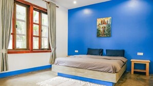 A bedroom with a bed, a small bedside table, a window with curtains, and a painting on the wall. - Last Summer Hostel & Bar (Siem Reap)