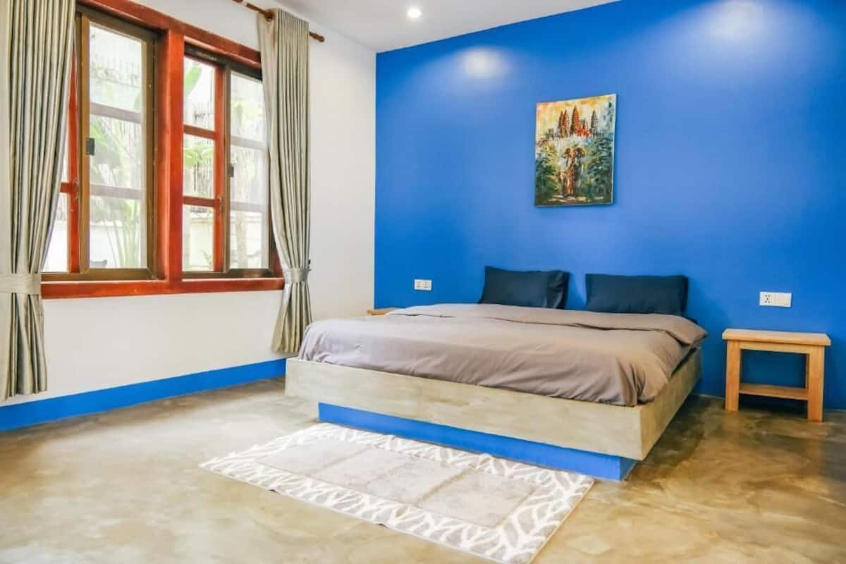 A bedroom with a bed, a small bedside table, a window with curtains, and a painting on the wall.