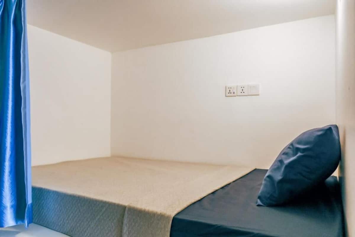 A small, single-bed room with a plain white wall, a power outlet, and a blue curtain.