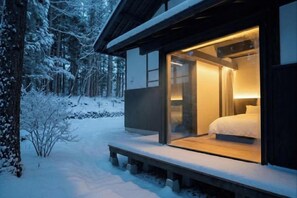 Une cabane moderne avec une entrée dotée d’une porte en verre, une terrasse en bois et un lit visible à l’intérieur.