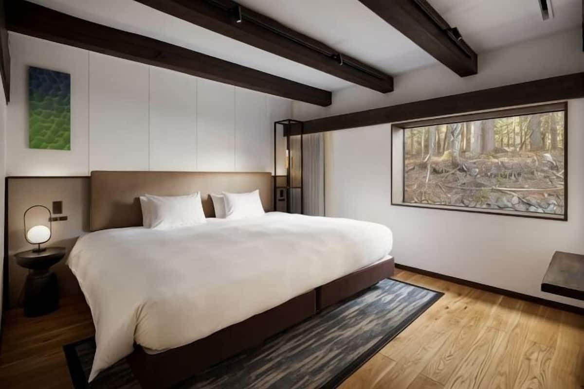 Una cama grande con cabecera, una mesita de noche con lámpara, un ventanal con vistas al bosque y suelo de madera.