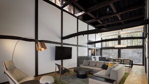 Una sala moderna con sofá, sillón y mesa de centro, un televisor de pantalla plana en la pared, y ventanas amplias que ofrecen vistas al exterior.