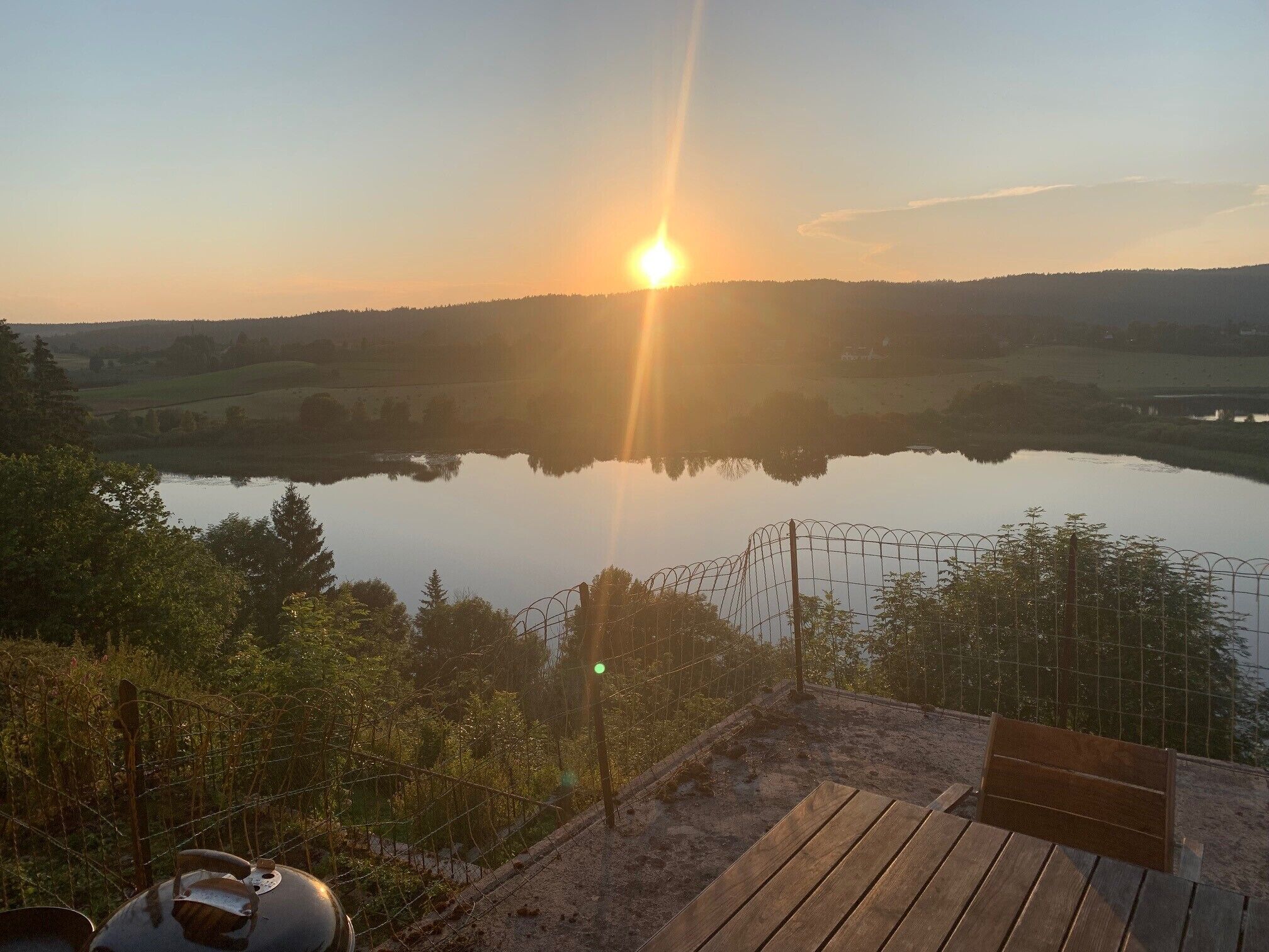 A sunset over a lake with a wooden deck and a metal fence in the foreground.