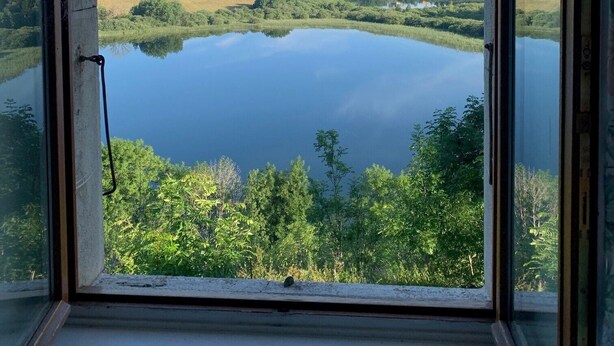 Vista a través de una ventana de un lago rodeado de vegetación y casas a lo lejos.