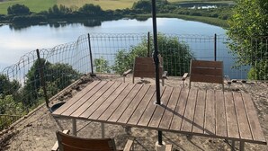 Una terraza de madera con una sombrilla grande, dos bancos de madera y vistas a un lago y colinas.