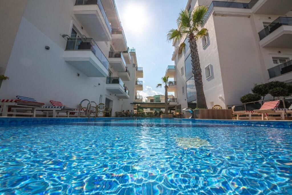 A swimming pool with clear blue water, surrounded by white buildings with balconies and palm trees.