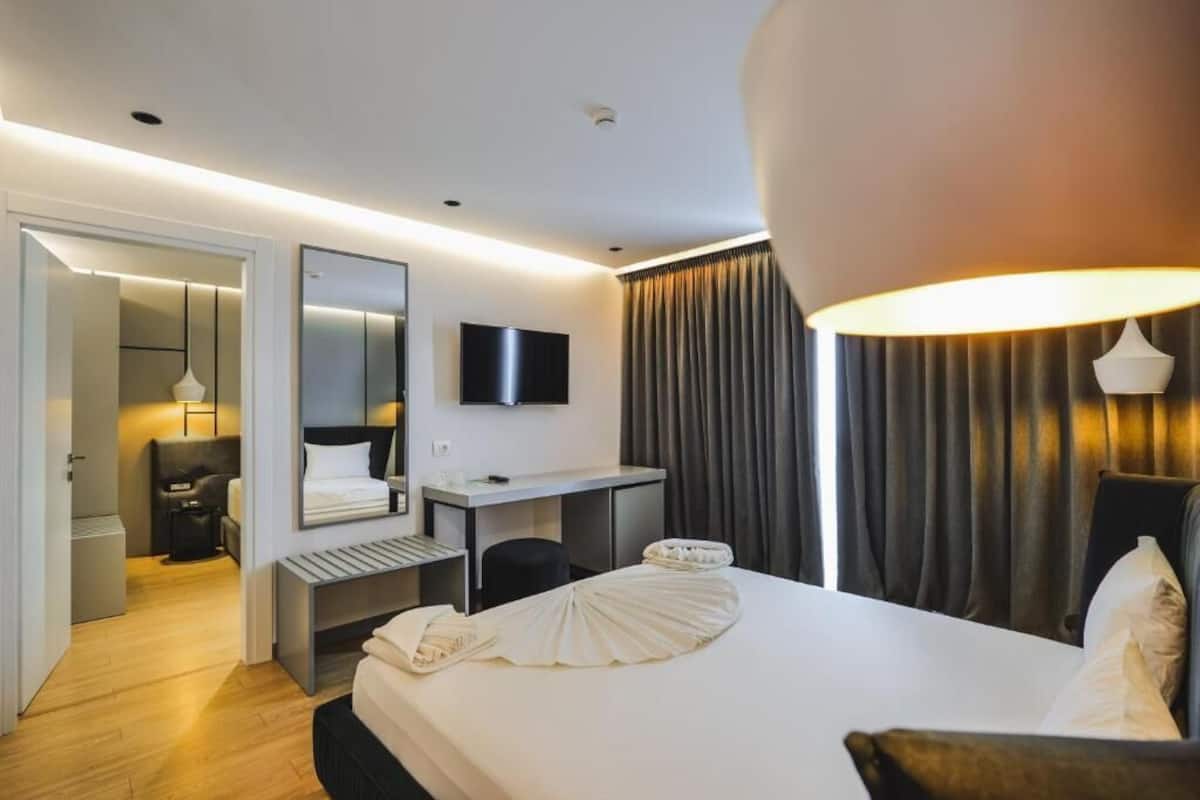 Una habitación de hotel con cama, escritorio, silla, televisor y un amplio ventanal con cortinas.
