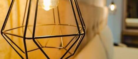 Una lámpara colgante de metal con diseño geométrico y bombilla visible, en una habitación con un sofá beige y una pared verde azulado al fondo.