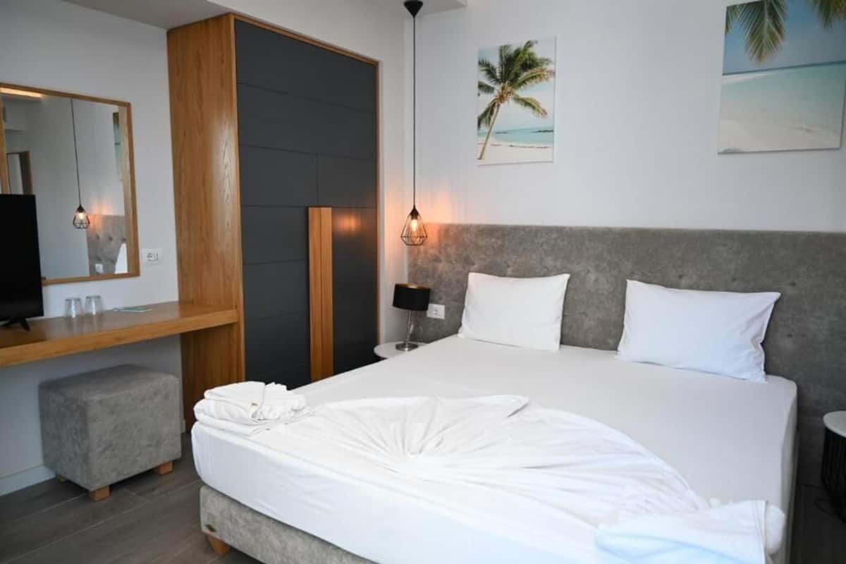 Una cama bien hecha con sábanas blancas, una mesita de noche de madera con una lámpara, un otoman gris y un espejo grande en la pared.