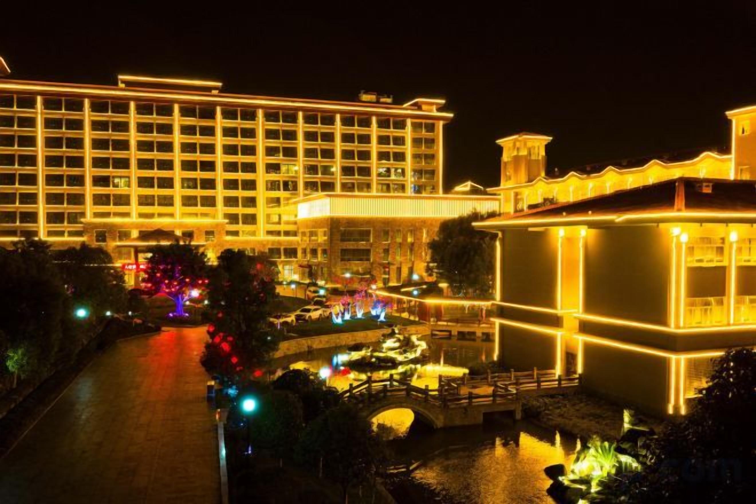 Un hotel grande con varios pisos y una fachada bien iluminada, un patio con una fuente y un puente que conecta diferentes áreas de la propiedad.
