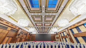 A large conference room with a stage, rows of chairs, and a high ceiling with chandeliers.