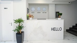 A modern reception desk with a "HELLO" sign, a clock displaying times for Tirana, London, and New York, and a potted plant.