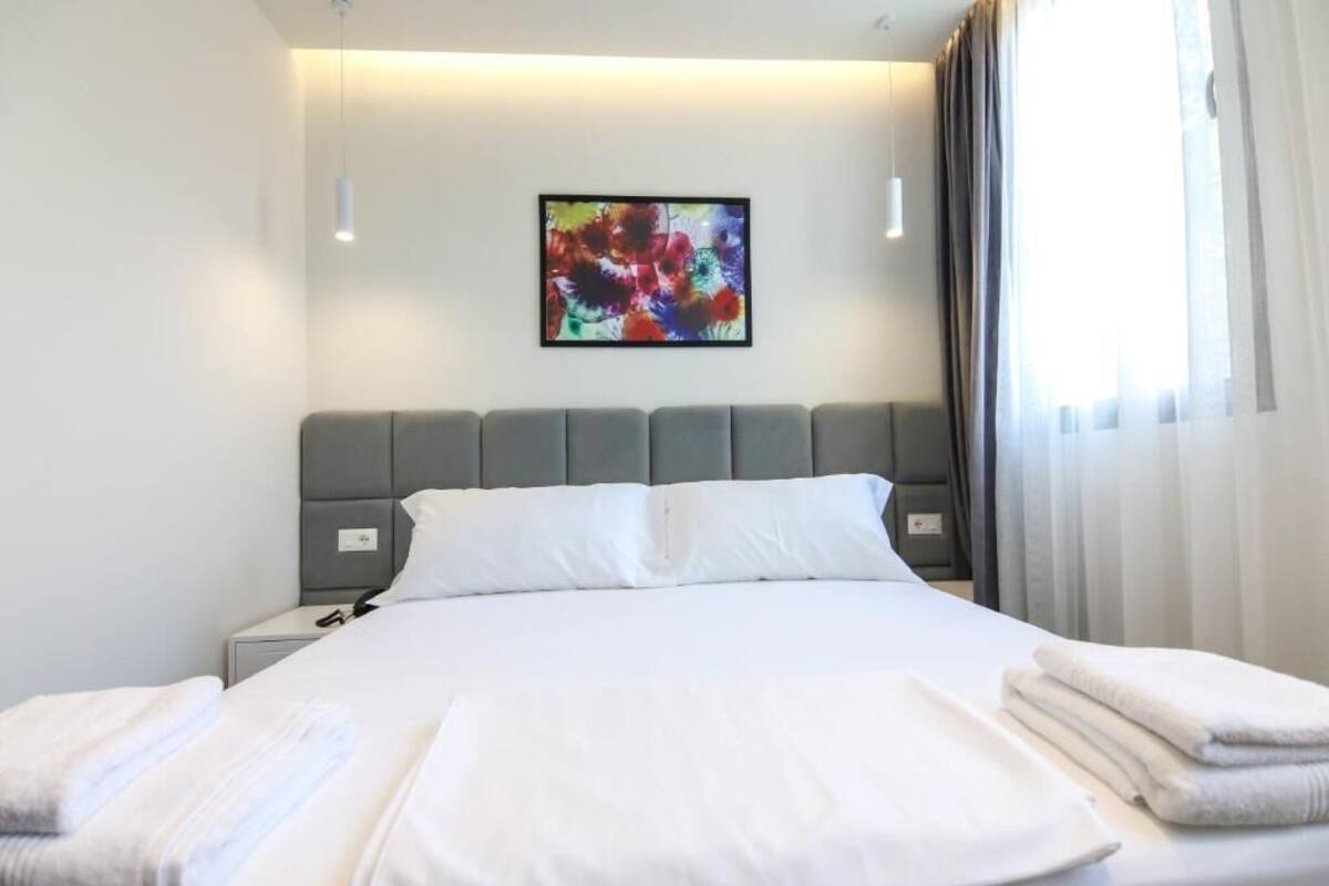 A hotel room with a bed, white linens, a gray headboard, a nightstand, a wall-mounted abstract painting, and a window with curtains.