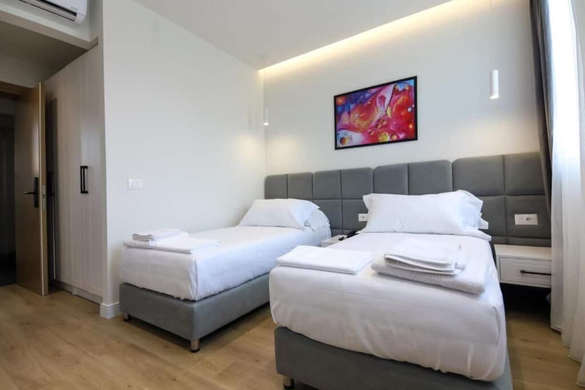 A hotel room with two beds, a gray headboard, white bedding, a colorful abstract painting, and a wooden floor.
