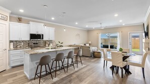 A modern kitchen with white cabinets, a central island with bar stools, a dining area with a table and chairs, and a view of the outdoors through a glass door.