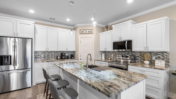 A modern kitchen with white cabinets, stainless steel appliances, a marble island, and a 'let's eat' sign.