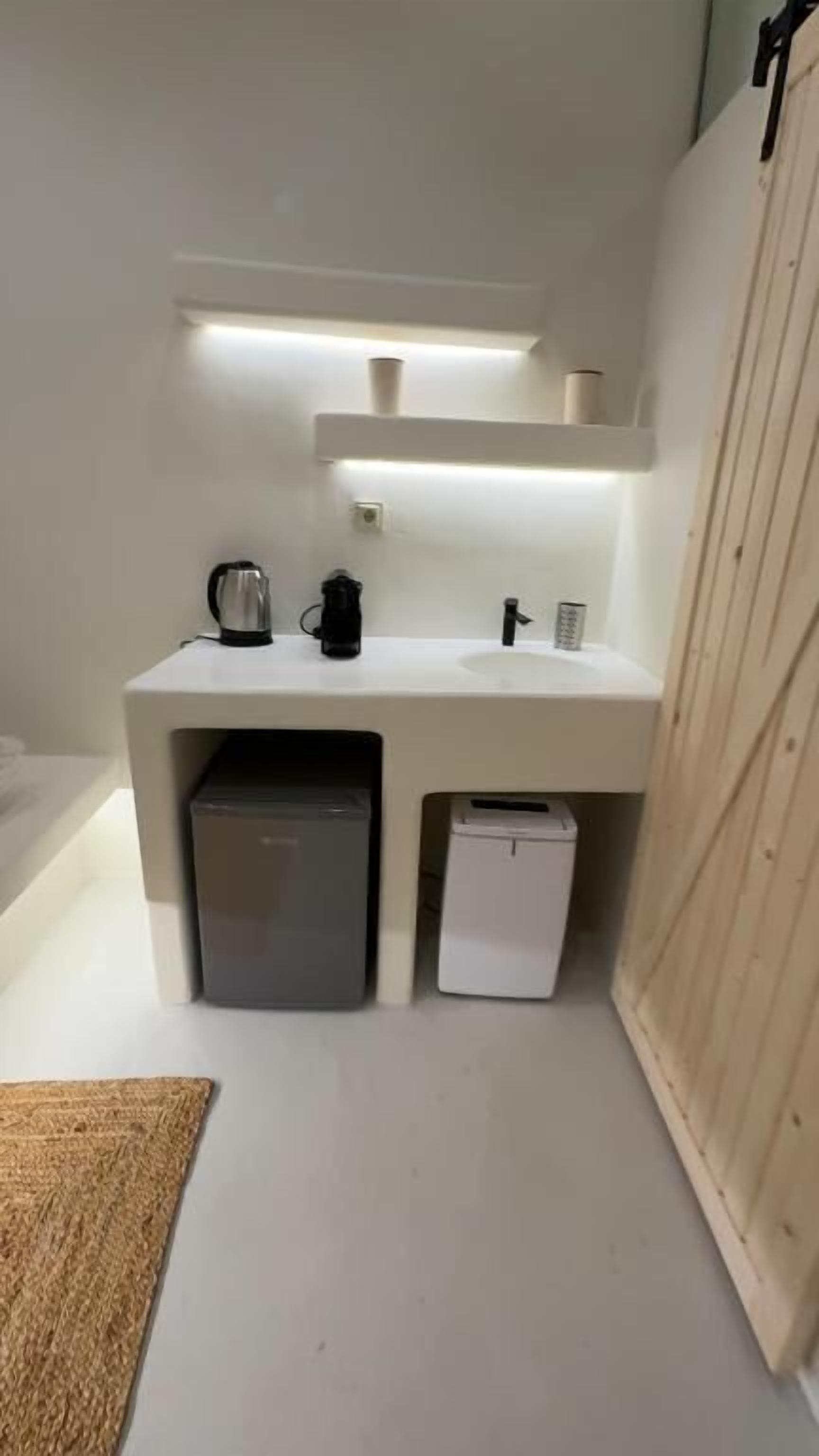 Una kitchenette compacta con una encimera blanca, cafetera, hervidor de agua y un refrigerador pequeño.