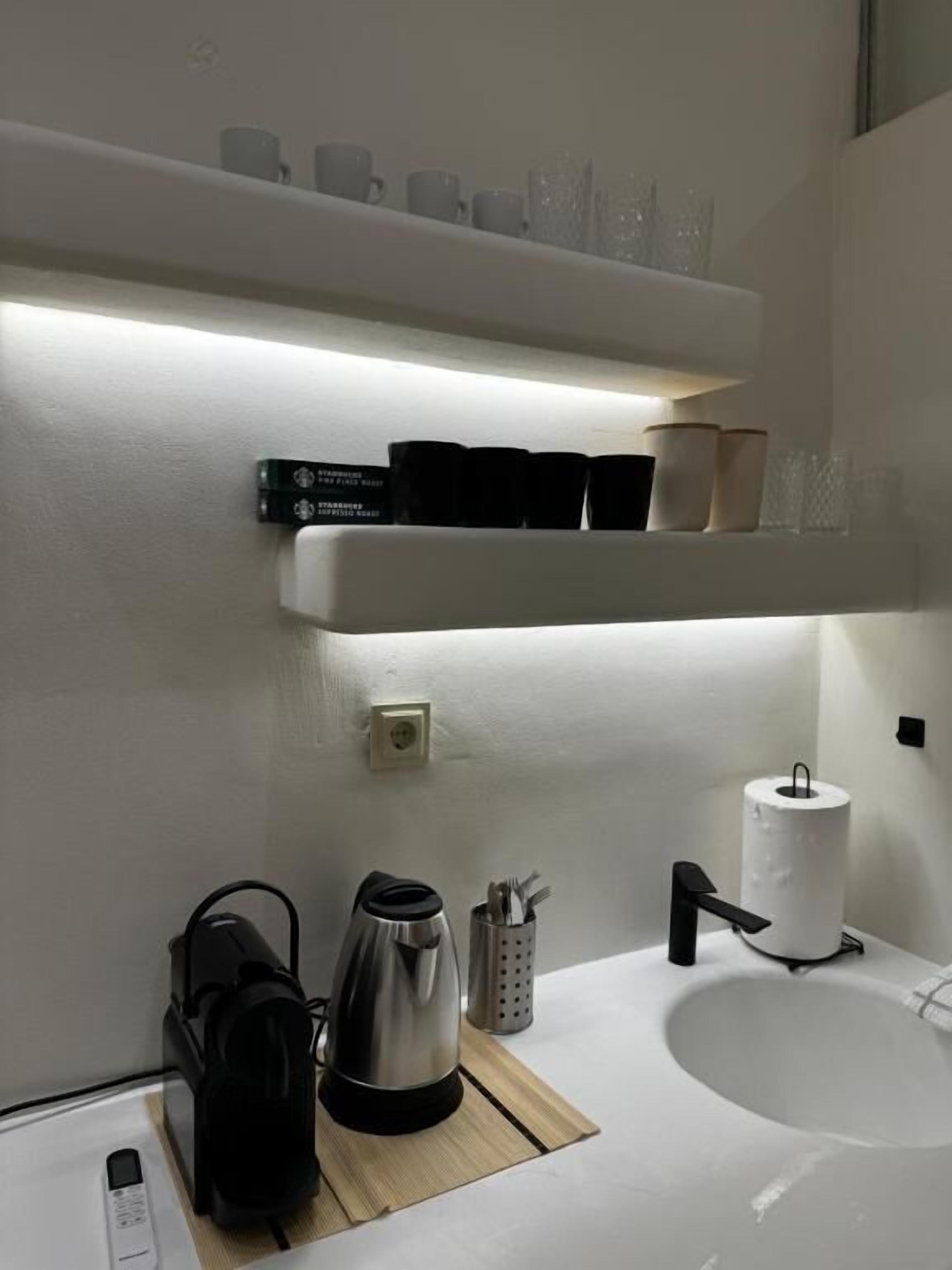 A modern bathroom with a white sink, a kettle, a coffee maker, and a shelf with cups and a box of tissues.