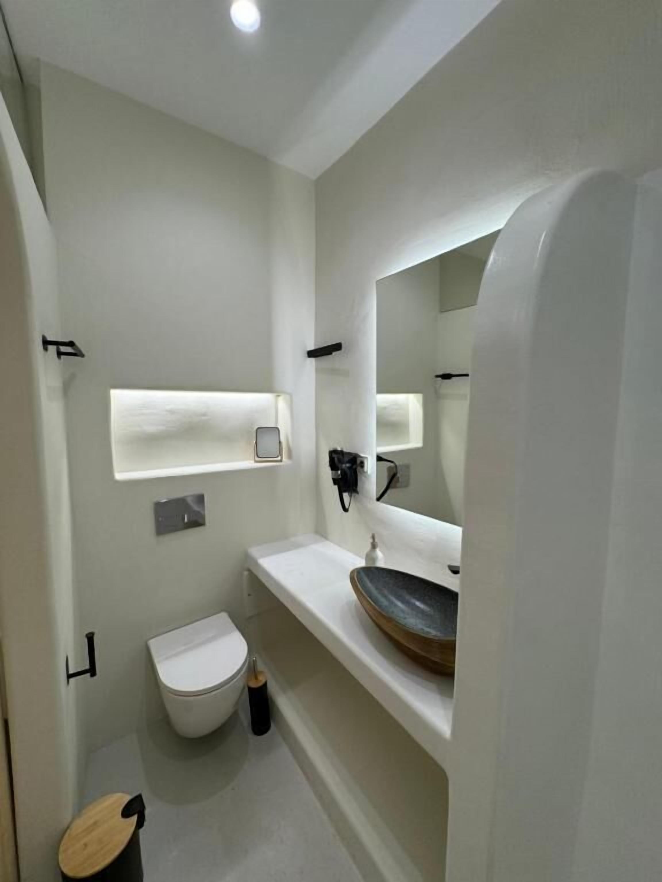 Une salle de bain moderne avec des toilettes blanches, un lavabo rond doté d’un plan de toilette en bois, et un miroir au-dessus du lavabo.