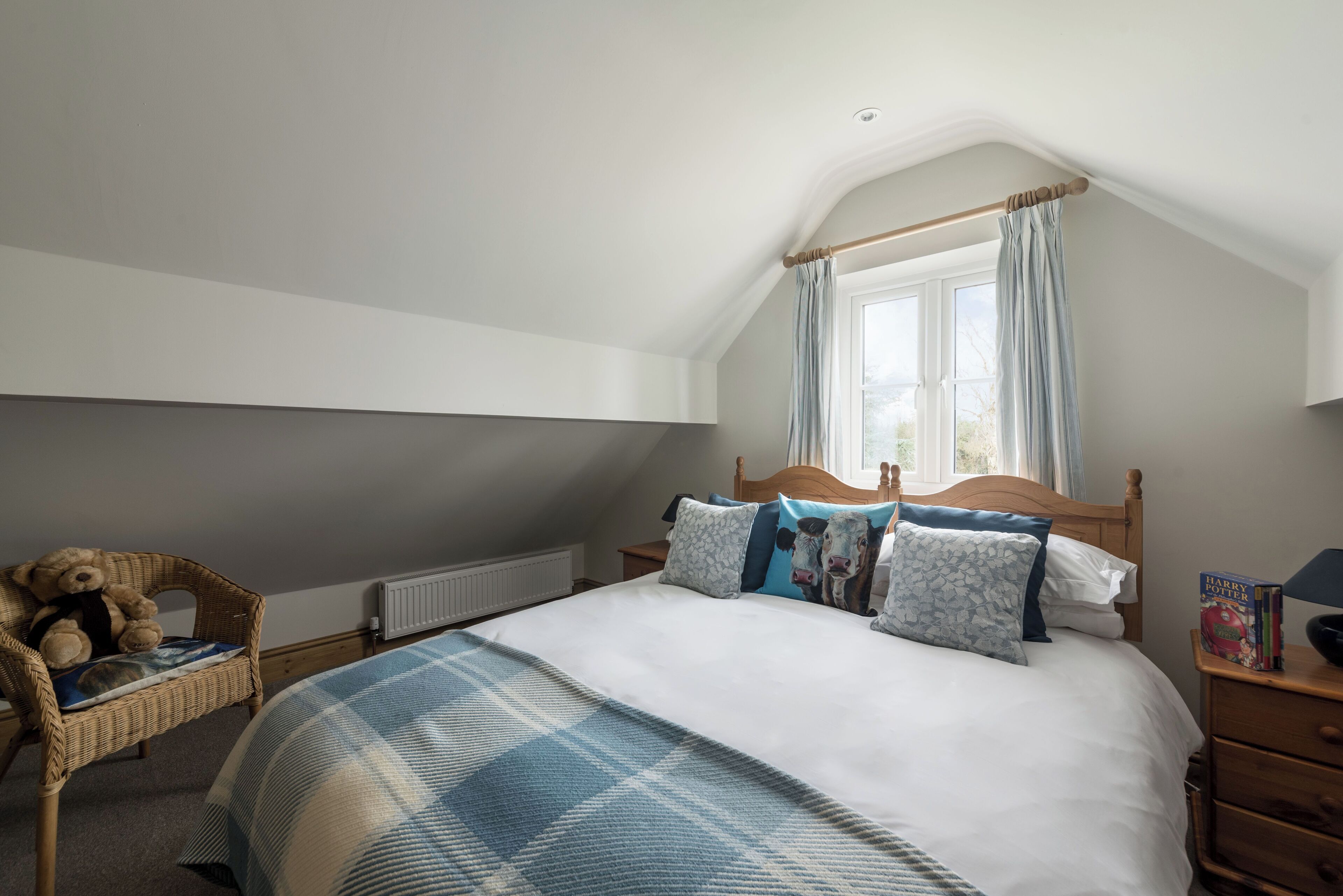 Een slaapkamer met een groot bed, een houten hoofdbord, een raam met gordijnen, een rieten fauteuil en een nachtkastje met boeken.