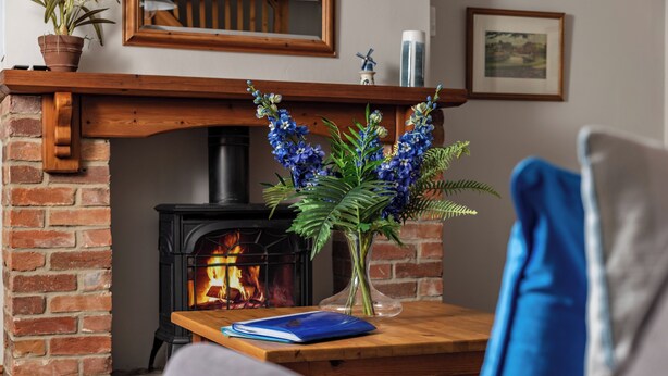 A cozy living room with a brick fireplace, a wooden table with a vase of flowers, and a blue book. - Swallow Cottage @ Olivemead Farm (Chippenham)