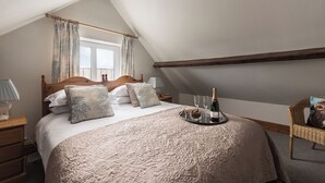 A bedroom with a large bed, a wooden headboard, a window with curtains, a nightstand with a lamp, and a tray with champagne glasses and a bottle. - Swallow Cottage @ Olivemead Farm (Chippenham)