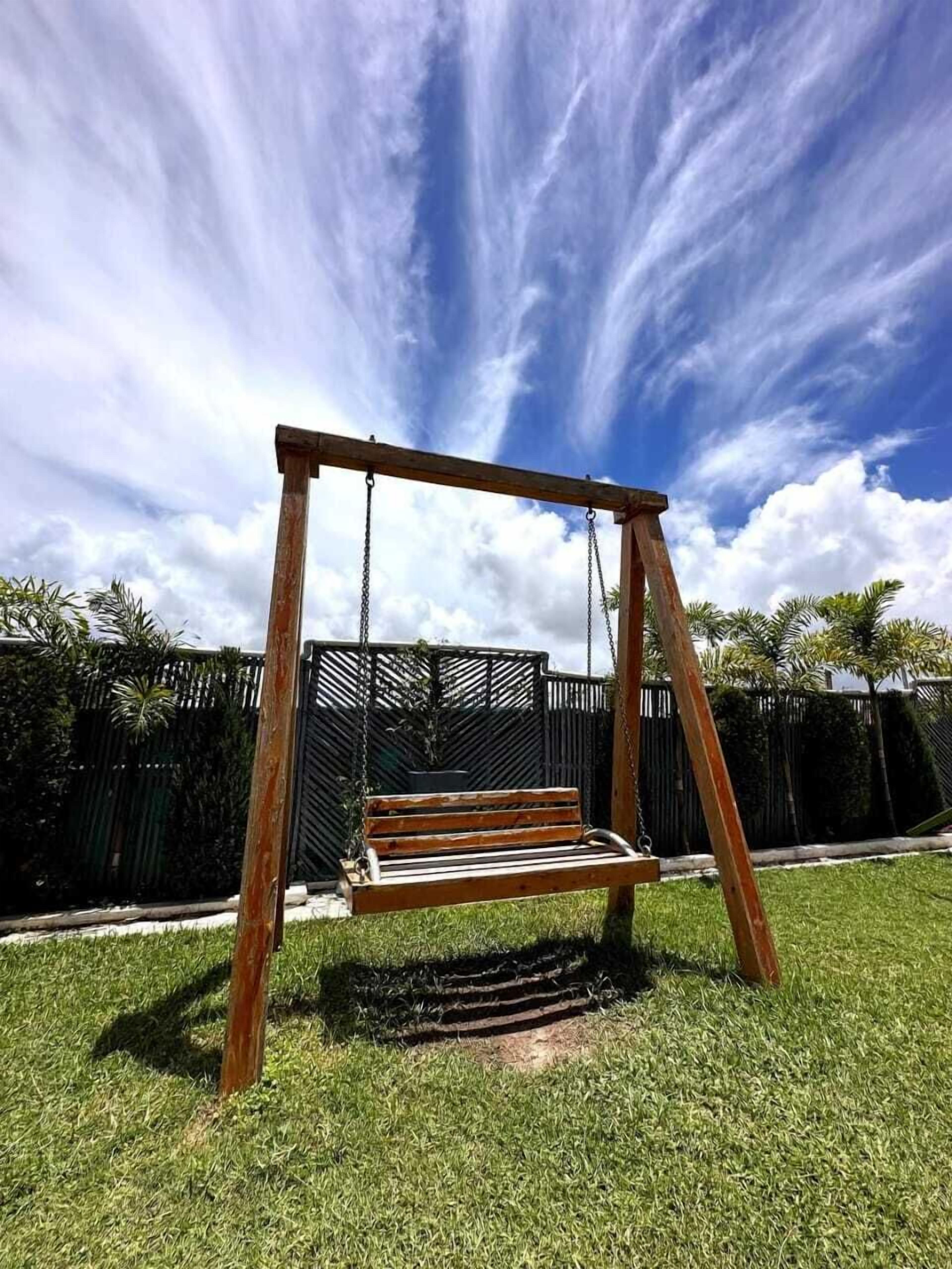 A wooden swing set in a grassy yard with a black fence and palm trees in the background.