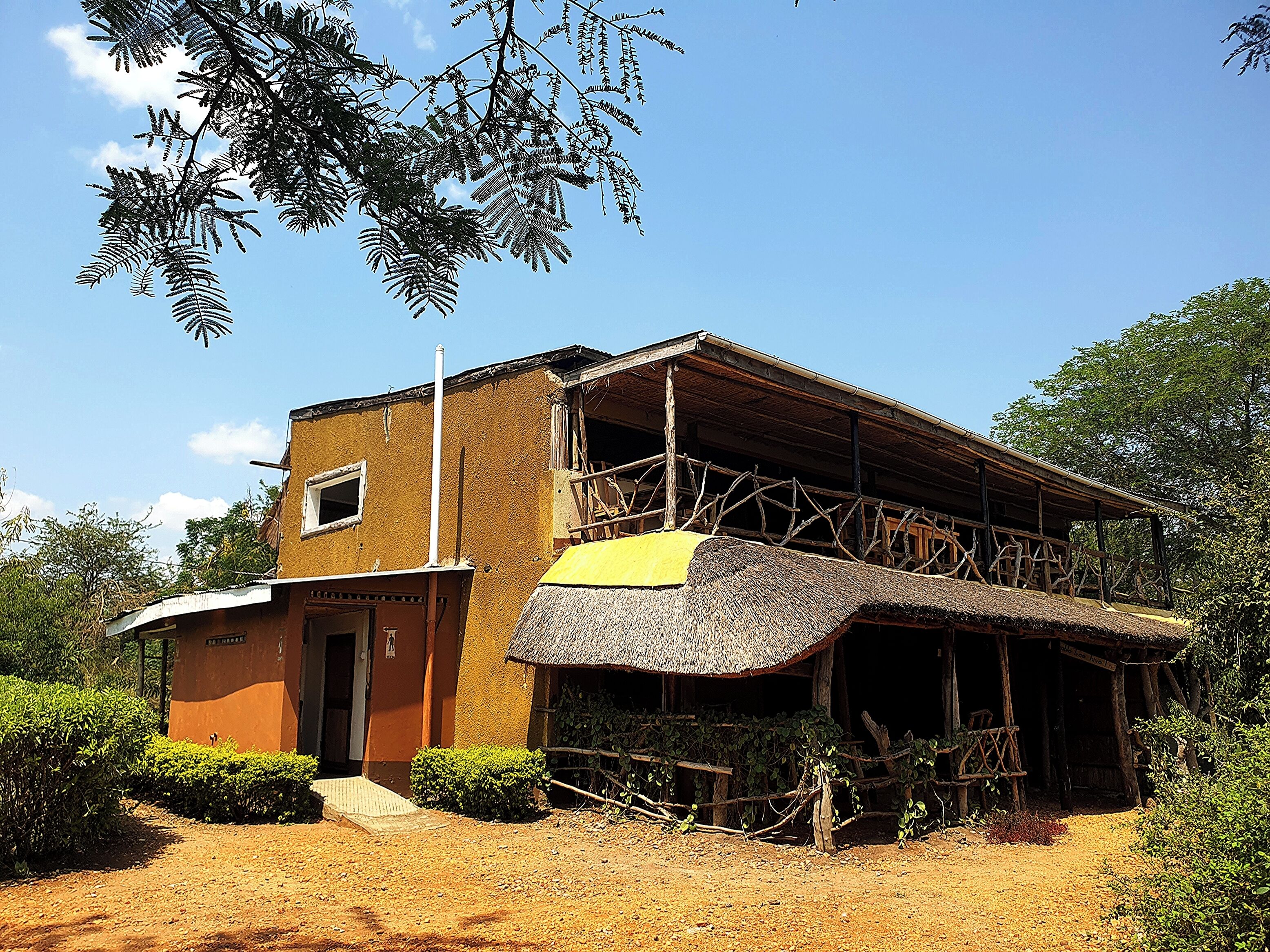 A rustic building with a thatched roof extension, surrounded by greenery.