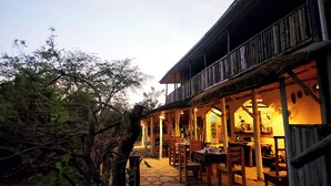 An outdoor dining area with stone flooring, wooden tables and chairs, and a wooden railing overlooking the space. - Hornbill Bush Lodge (Pakwach)