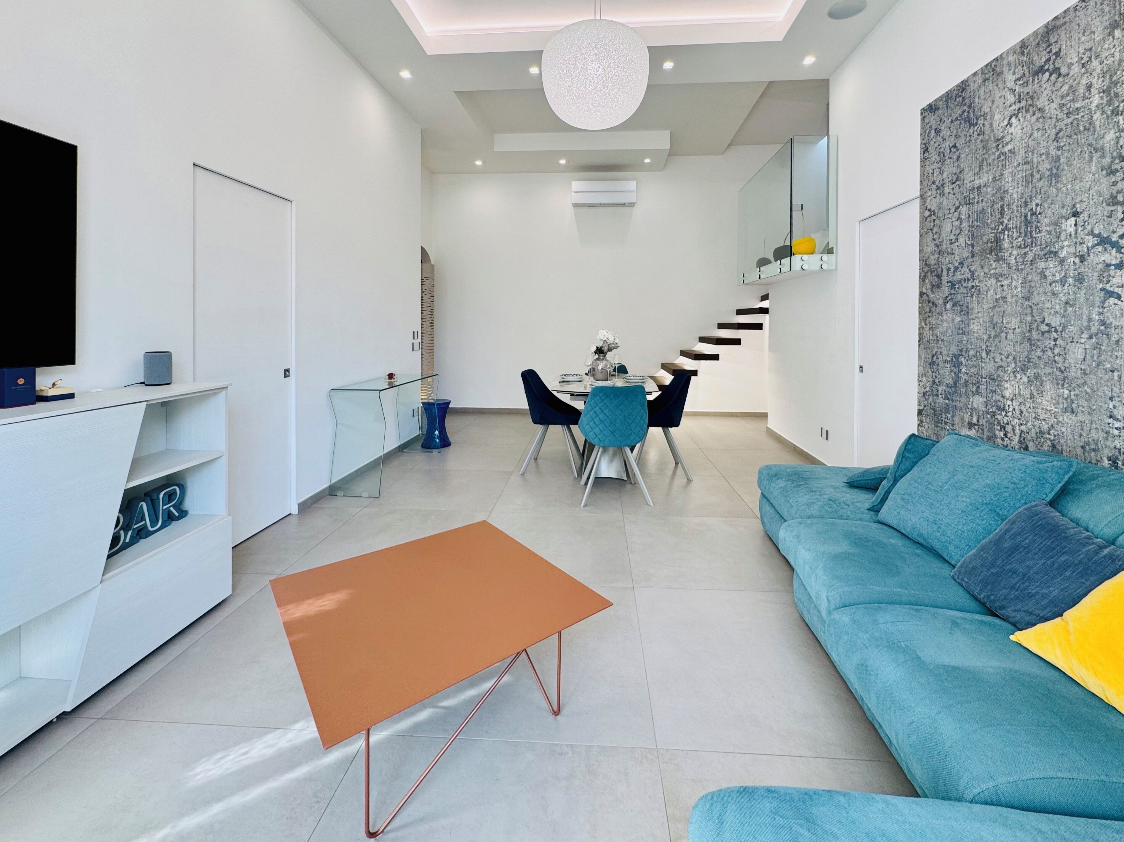 Uma sala de estar moderna com um sofá, uma mesa de centro, uma área de jantar com cadeiras, uma TV e uma escadaria para outro nível.