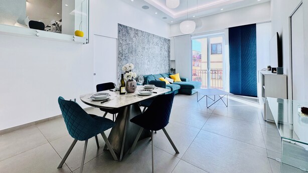 A modern living room with a sofa, a dining table with chairs, and a large window. - YourHome - AC Luxury House (Meta)
