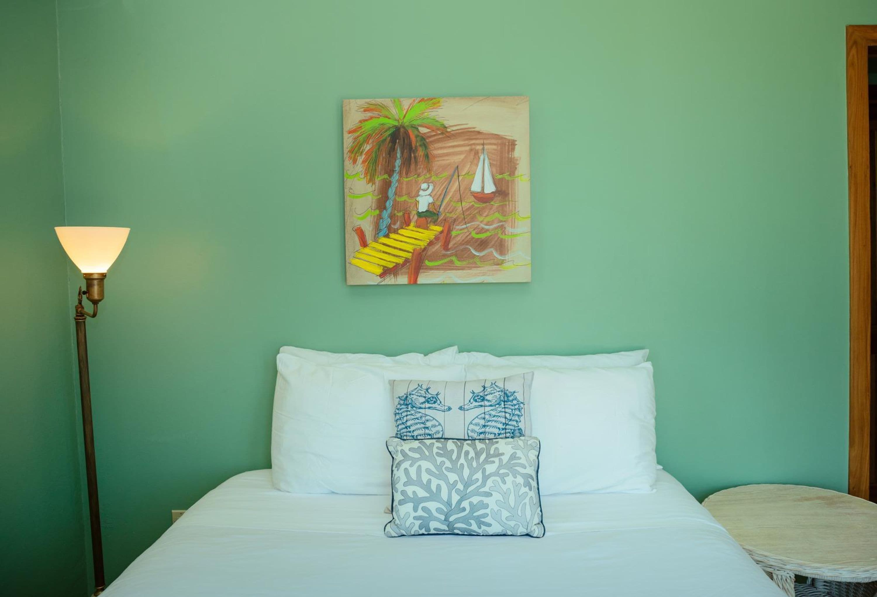 Una cama con sábanas blancas, dos almohadas decorativas, un cuadro de una escena tropical y una lámpara de pie.