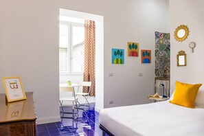 Une chambre d’hôtel avec un lit, un bureau et une chaise. Les murs sont ornés de tableaux et il y a une fenêtre avec des rideaux.