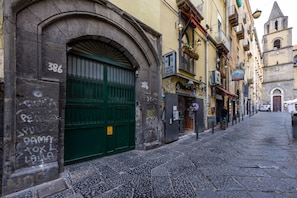 Une rue étroite avec une porte verte et un arc en pierre, des bâtiments dotés de balcons et, au loin, le clocher d’une église.