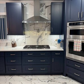 A modern kitchen with dark blue cabinets, a stainless steel range hood, a built-in oven, and marble countertops.