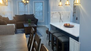 A modern living room with a sofa, dining table, and chairs. There is a kitchen area with a counter and stools.