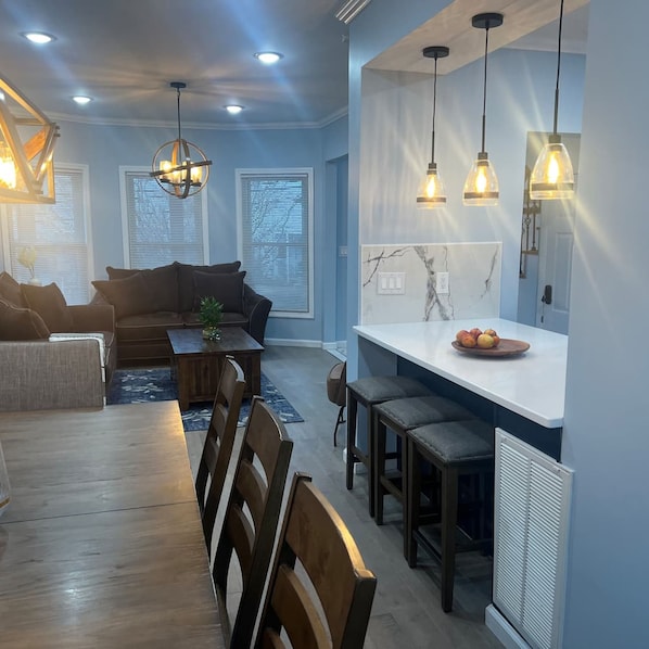 A modern living room with a sofa, dining table, and chairs. There is a kitchen area with a counter and stools.