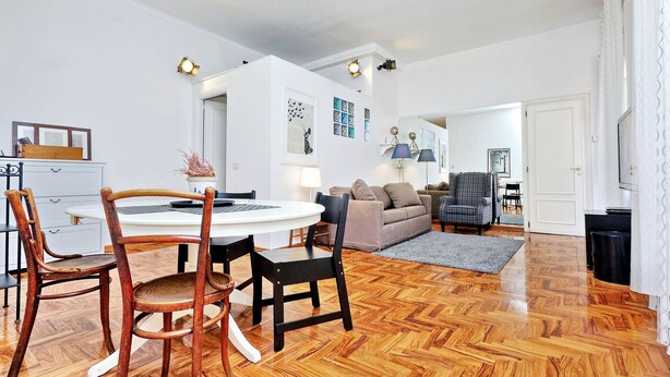 A modern living room with a round dining table, two wooden chairs, a sofa, and a grey armchair. - 4bnb - Lante Apartment (Rome)