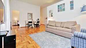 A modern living room with a wooden floor, a beige sofa, a blue plaid armchair, and a dining area with a table and chairs. - 4bnb - Lante Apartment (Rome)