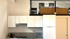 Een kleine keuken met witte kasten, een roestvrijstalen spoelbak en een klein aanrecht. Er staat een eettafel met vier stoelen en een potplant erop.