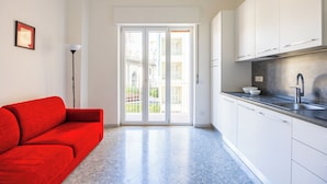 Una cocina moderna con armarios blancos, un lavamanos de acero inoxidable y un sofá rojo en una habitación luminosa con un amplio ventanal corredera que da a un balcón exterior.
