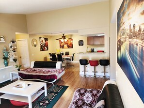 Un salon avec un canapé, une table à manger et des chaises, un coin cuisine et un grand tableau accroché au mur.