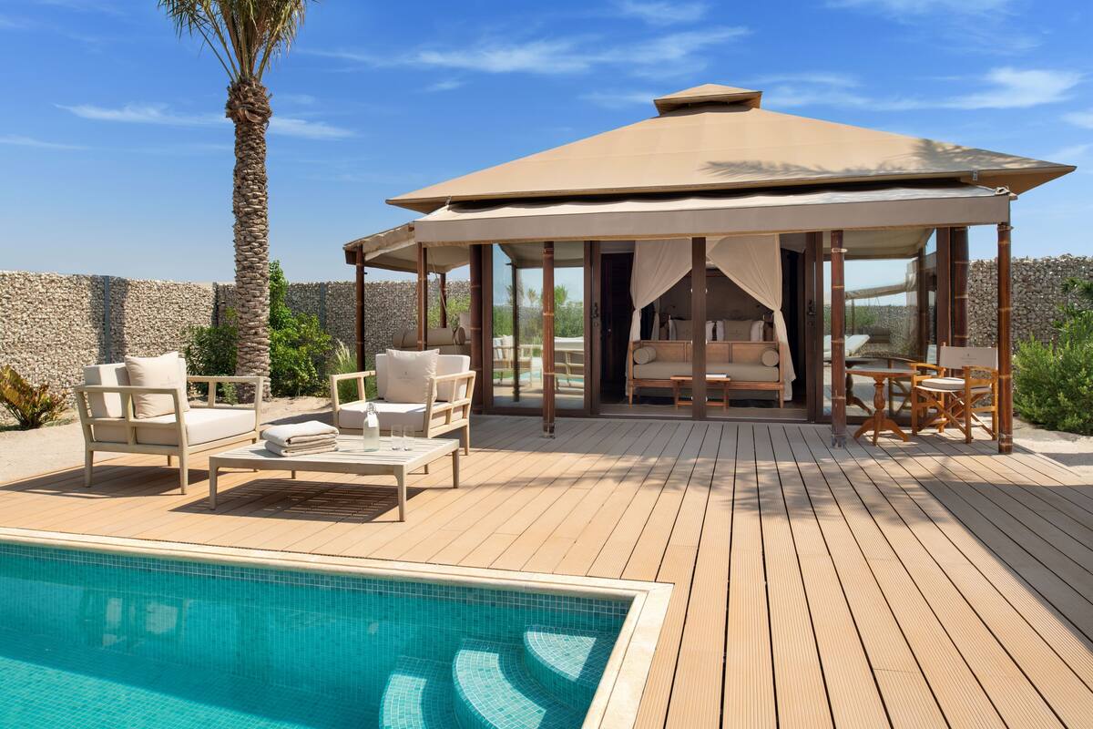 Zona de piscina con un espacio de estar techado, sillas de descanso y una terraza de madera.