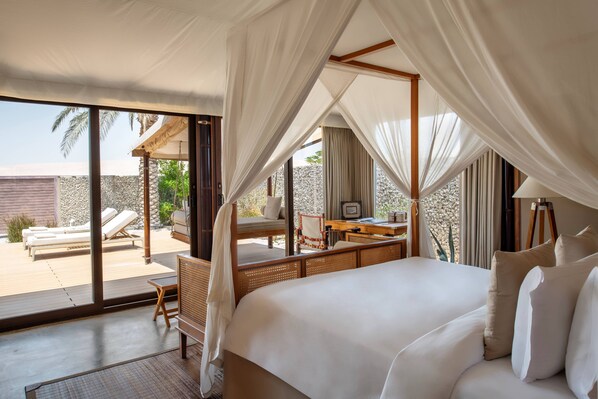 A bedroom with a large bed, a desk, and a chair. There is a balcony with lounge chairs and a view of the outdoors. - The Outpost Al Barari (Mesaieed)