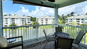 Un balcon abrité, doté d’un ventilateur de plafond, de deux chaises, d’une petite table et offrant une vue sur un lac et des bâtiments en arrière-plan.