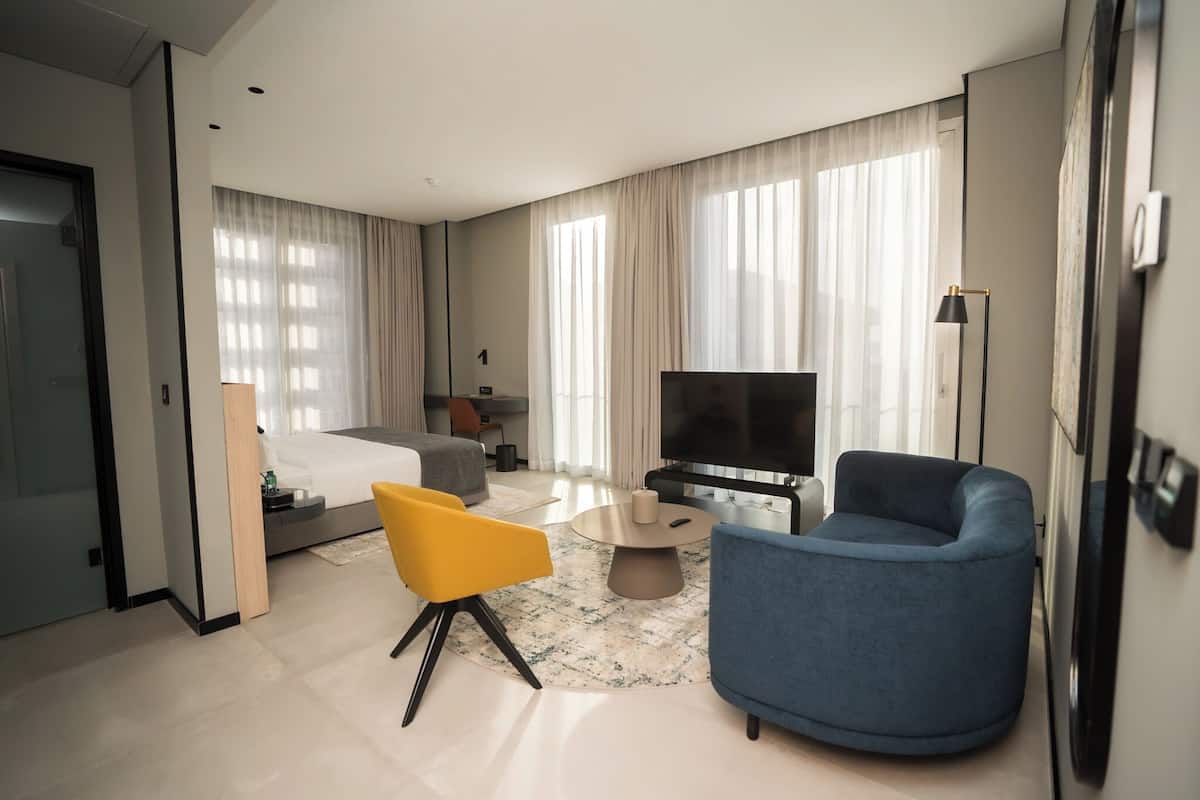 A modern hotel room with a bed, a TV, a sofa, a chair, and a coffee table.