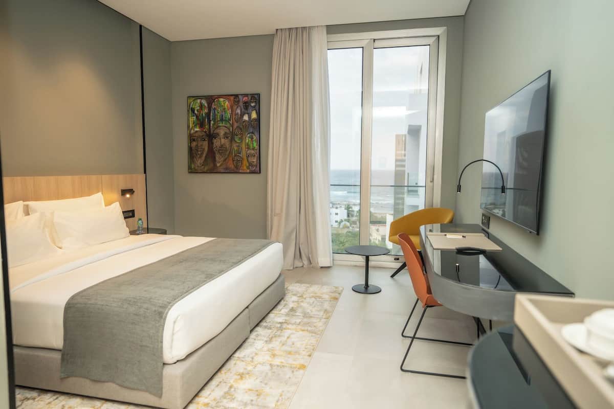 A hotel room with a bed, a desk, a chair, a TV, a painting, and a view of the city.