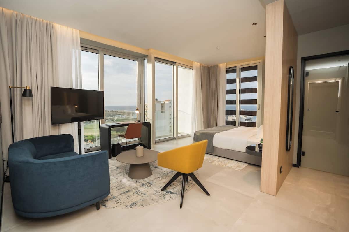 A modern hotel room with a large window, a flat-screen TV, a sofa, a yellow chair, and a bed.
