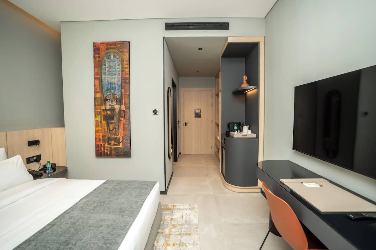 A modern hotel room with a bed, desk, TV, and artwork on the wall.