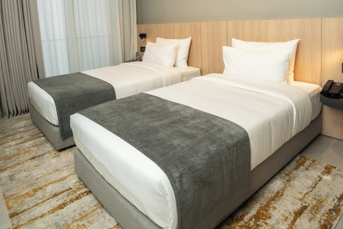 A hotel room with two beds, a wooden headboard, a nightstand, and a large window with curtains.